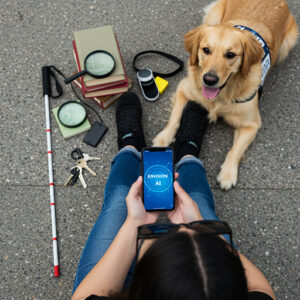Osoba sedí na zemi s&nbsp;nohama nataženýma před sebou. V&nbsp;rukou drží mobilní telefon, na kterém je zobrazeno "ENVISION AI". Vedle osoby leží vodicí pes, pravděpodobně zlatý retrívr, s&nbsp;postrojem. Na zemi vedle osoby jsou položeny různé předměty: bílá hůl s&nbsp;červenými pruhy, klíče, černý obal na telefon, dvě lupy, několik knih, černý popruh a&nbsp;malý bílý a&nbsp;černý přístroj. Pes má vyplazený jazyk a&nbsp;vypadá spokojeně. Osoba má na sobě modré džíny a&nbsp;černé boty. Vytvořeno pomocí AI.