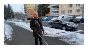 Na chodníku v sídlišti jde s bílou holí Linda, na krku má nasazen přístroj Biped NOA. Na trávníku je sníh.