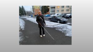 Na chodníku v sídlišti jde s bílou holí Linda, na krku má nasazen přístroj Biped NOA. Na trávníku je sníh.